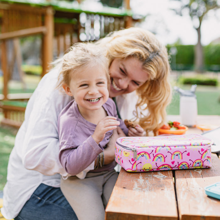 A smiling child sits on a woman’s lap at an outdoor table, with the Nuby Insulated Bento Box Lunch Box | Stars nearby and a playground in the background. Both appear happy, enjoying their time together.