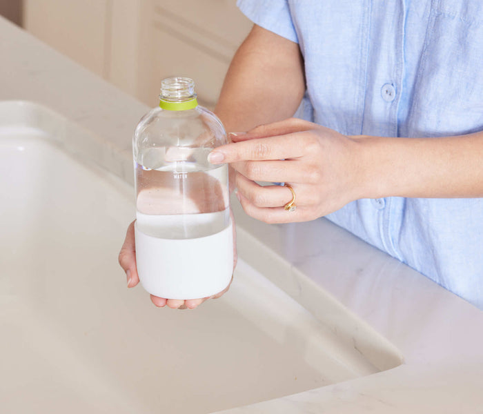 A woman standing at a kitchen faucet, pointing at the fill-lines on a refillable Glass Branch Basics bottle; there are separate fill-lines for water and Branch Basics Concentrate, respectively.