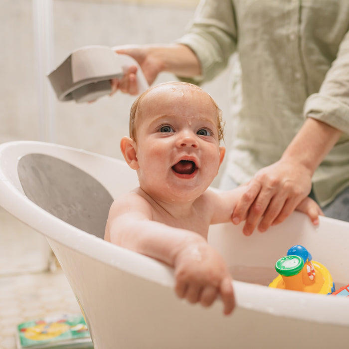 A smiling baby sits in a white tub filled with water and colorful toys, supported by an adult using the Baby Bathtime Essentials Bundle, which includes a safety spout guard and bathtub arm rest. The baby looks happy and excited.