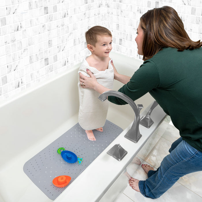 A woman kneels by a bathtub, wrapping a smiling child in a towel. The child stands on the Nuby Cushioned Bath Mat | Grey, with two toy boats nearby. The bathroom features white tile walls and a light floor.