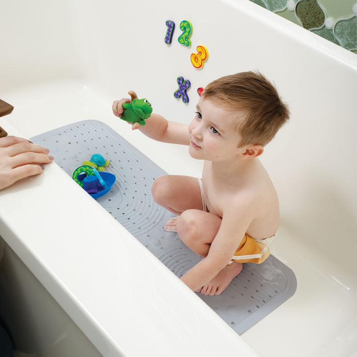 A young child crouches on a Nuby Cushioned Bath Mat | Grey in the tub, holding a green toy frog. Colorful numbers and an X stick to the wall, with an adult’s hand at the edge.