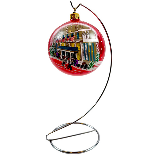A red Christmas ornament displayed on a curved metal stand against a plain white background. The ornament features a hand-painted winter city scene with snow, trees, and colorful buildings.