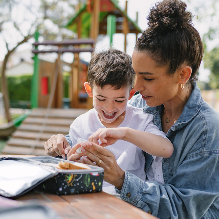 A woman and a young boy smile as they open the Nuby Insulated Bento Box Lunch Box | Stars at an outdoor picnic table, with playground equipment and sunshine in the background.