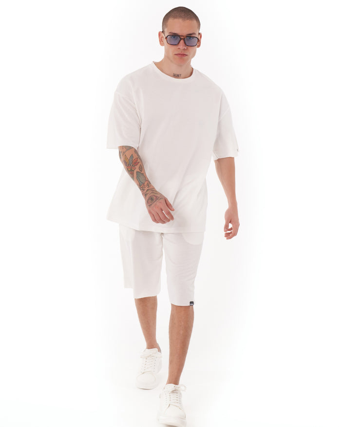 Martin Valen men's oversized white shortsuit, three-quarter front view of model walking, wearing sunglasses and showing tattooed forearm