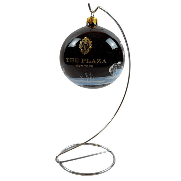 A spherical black Christmas ornament featuring a gold crest and the text 'THE PLAZA NEW YORK', suspended from a curved silver metal stand against a plain white background.