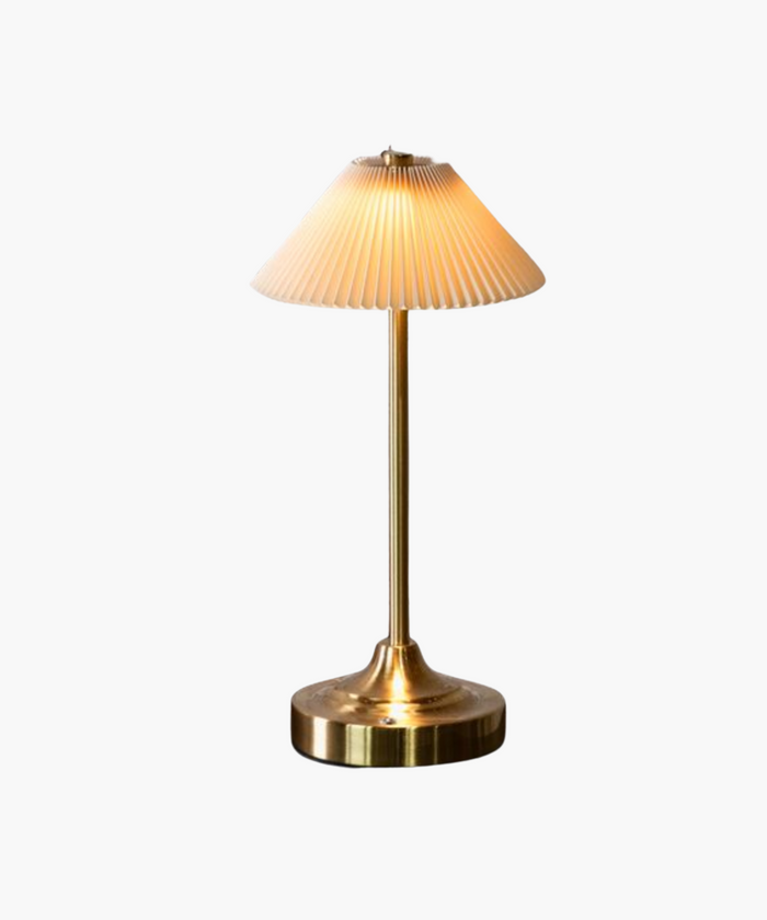 A warm, glowing brass table lamp with a fluted white shade sits on a reflective surface. The sleek design and soft lighting create an elegant and cozy ambiance. | Wildwood Home Co.