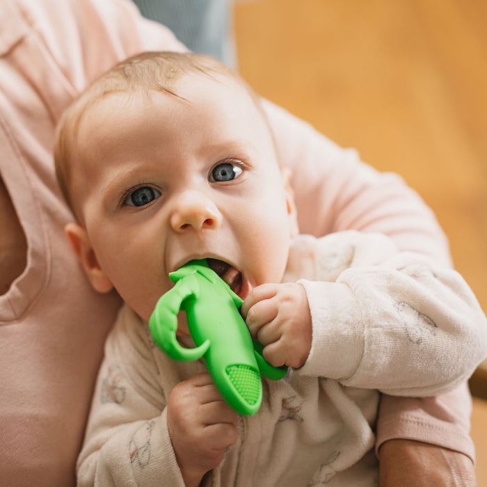 A baby in light pajamas chews on the Silicone Veggie Teether | Carrot for teething relief while being held by an adult, with a wooden floor in the background.